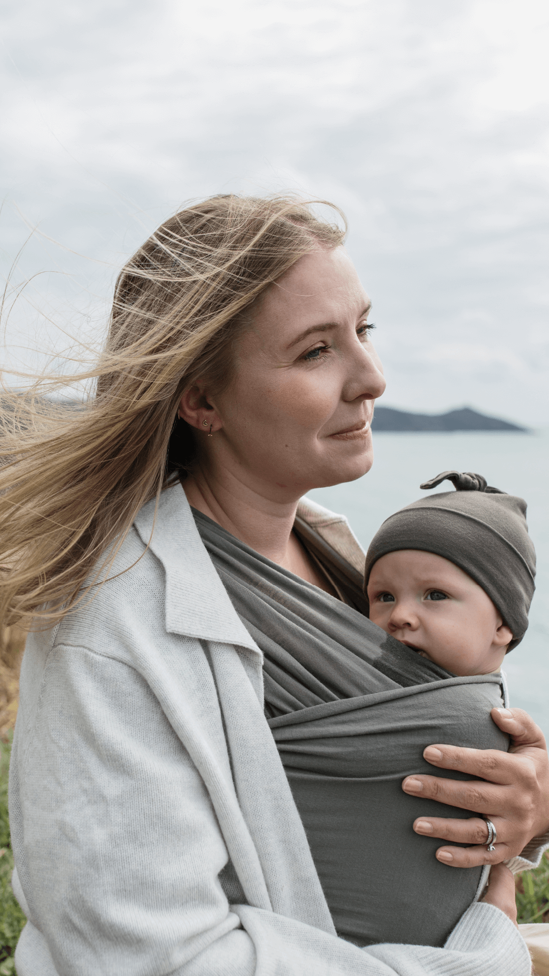 Mother's blond hair blows in the wind while holding baby in Sea Stone Solly Wrap