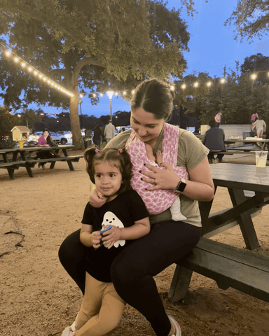 Mother sits on outdoor picnic table bench holding a baby in Ditsy Floral Solly Wrap with an arm around a young daughter