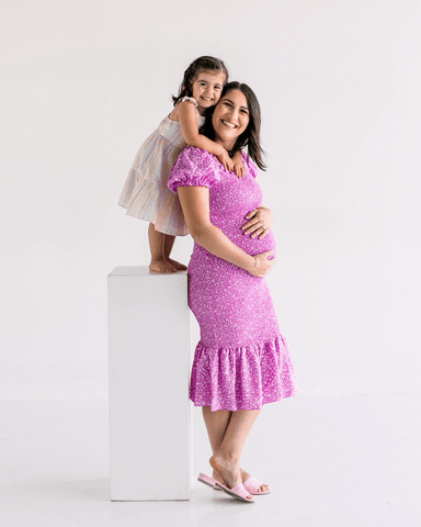 Pregnant woman smiling and holding her belly while her young daughter hugs her shoulders from behind and smiles.