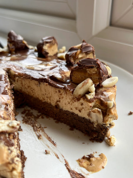 Vegan cheesecake with a slice cut out and layers visible. There is a biscuit base, the cheesecake, and a rich peanut butter topping.