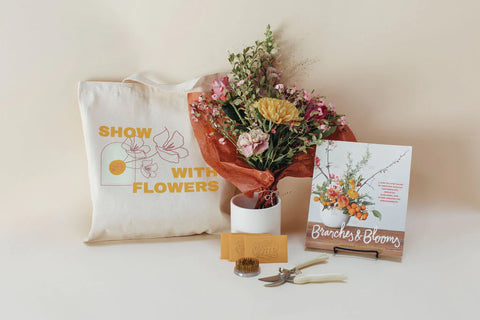 Image includes a tote with the saying "show up with flowers", a vase with brightly colored flowers arranged, a pair of shears, packets of flower food, a frog, and a guidebook for flower arranging