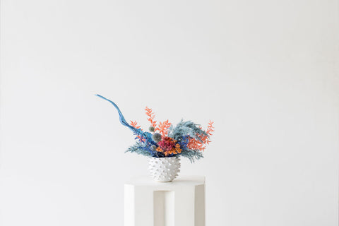 The coral reef everlasting arrangement adds a vibrant touch to any home decor with its bold painted dried ingredients, including pampas grass, banana leaf, thistle, and ruscus. The materials are carefully arranged and designed in a decorative ceramic vase, allowing your space to feel tropical and fashionable. 