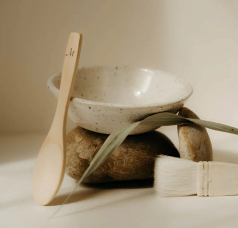 An image of a wooden spoon, ceramic bowl, goat-hair brush surrounded by rocks and a grass reed