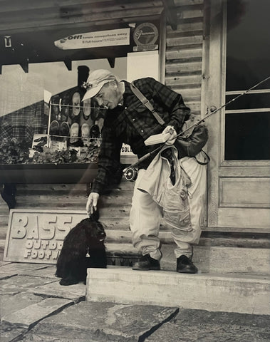 George Fletcher standing on the steps of the Rangeley Region Sport Shop, dressed for fly fishing and patting a black dog