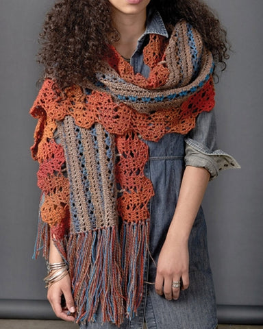 Woman standing wearing a funky bohemian crocheted long rectangular crocheted scarf with a blue and tan center section and along the long edges it features a orange wavy trim. The short ends are finished long tassels to create a fringed edge.