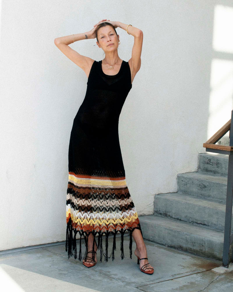 Model stands against a white wall wearing a black knitted dress with her arms above her head.