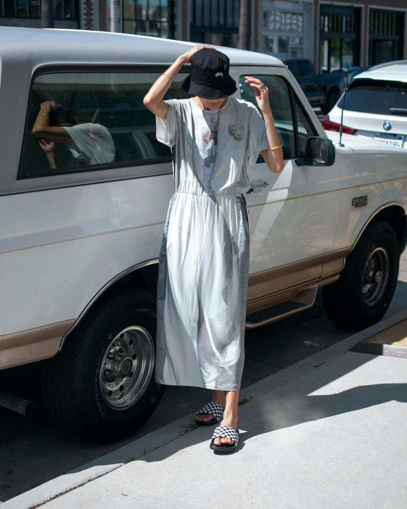 Model stands in front of a cream colored car, wearing a printed grey and white dress, black bucket hat, and checkered slide sandals.