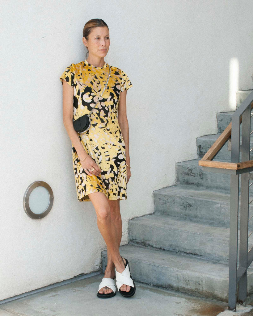 Model stands against white wall wearing a black and yellow printed mini dress and white sandals.