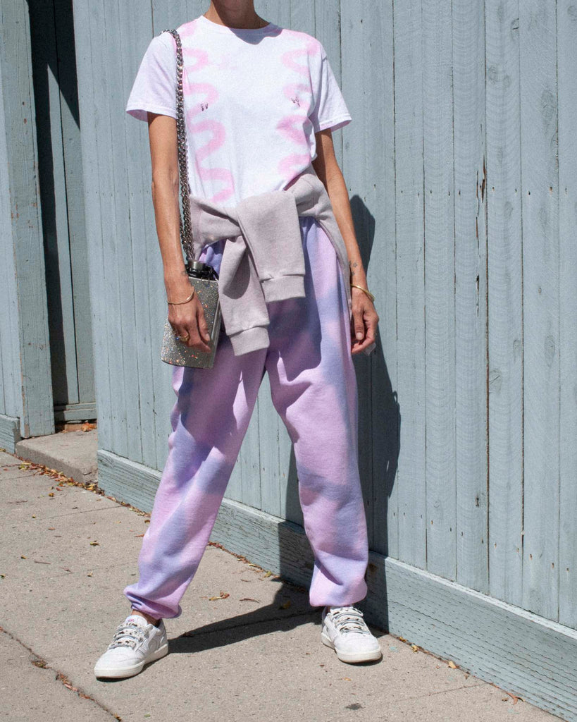 Model wears a pink and purple tie-dyed t-shirt and sweatpants on sidewalk in front of a blue wall.