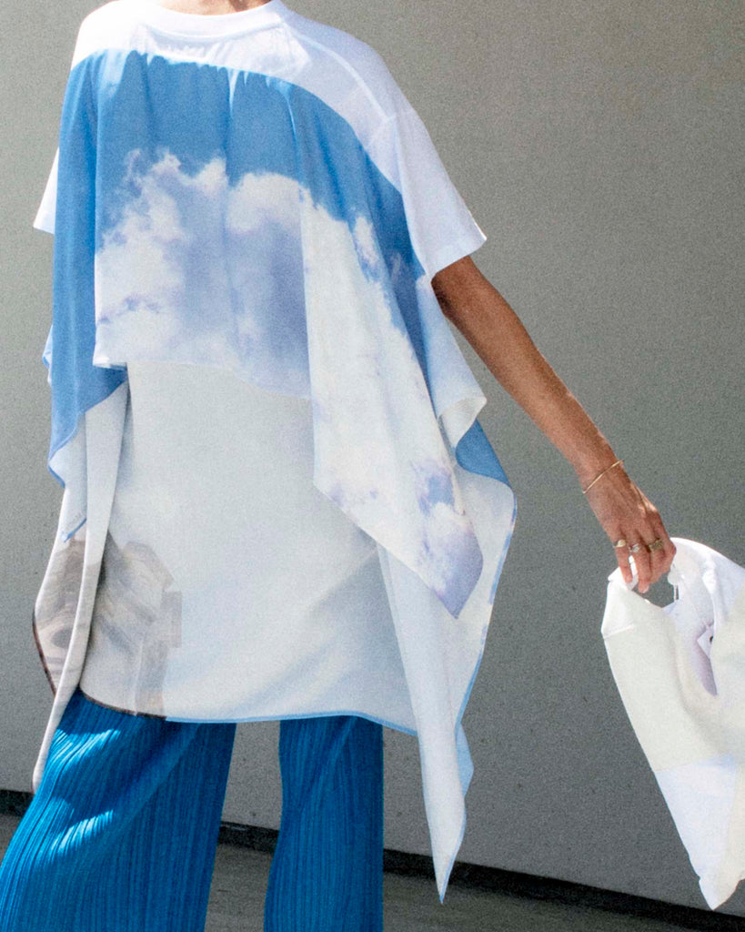 Back view of model wearing cloud print t-shirt and blue pants, holding white bag.