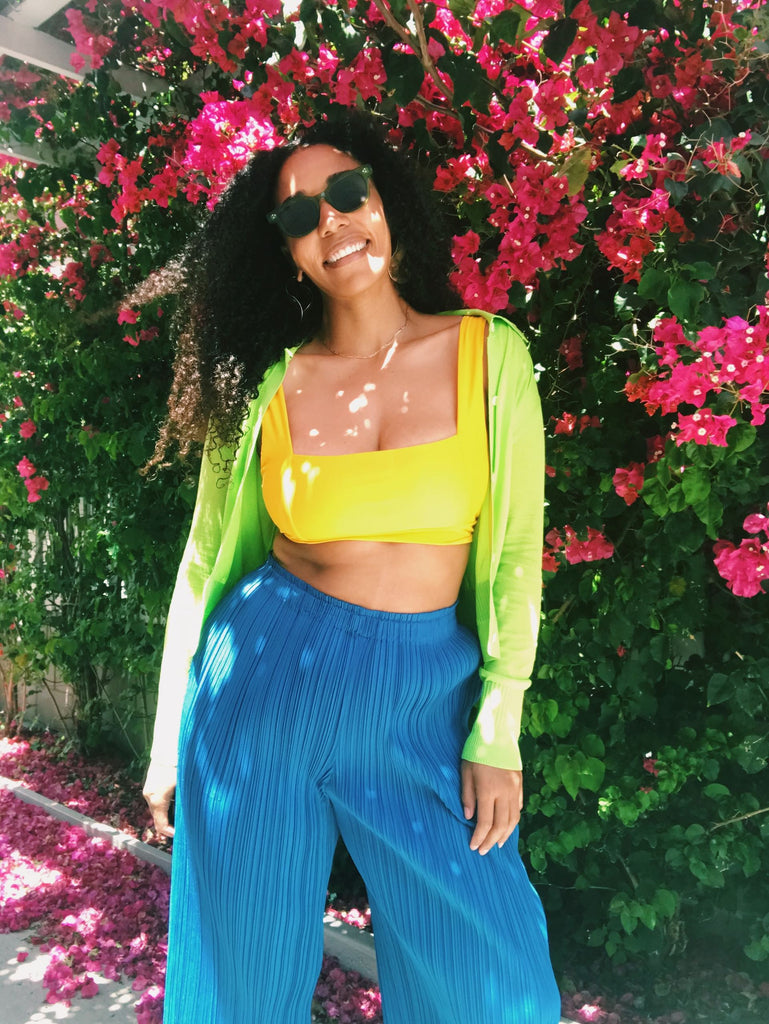 Model stands in front of bougainvillea, wearing a yellow sports bra, neon green cardigan, and electric blue pleated pants.
