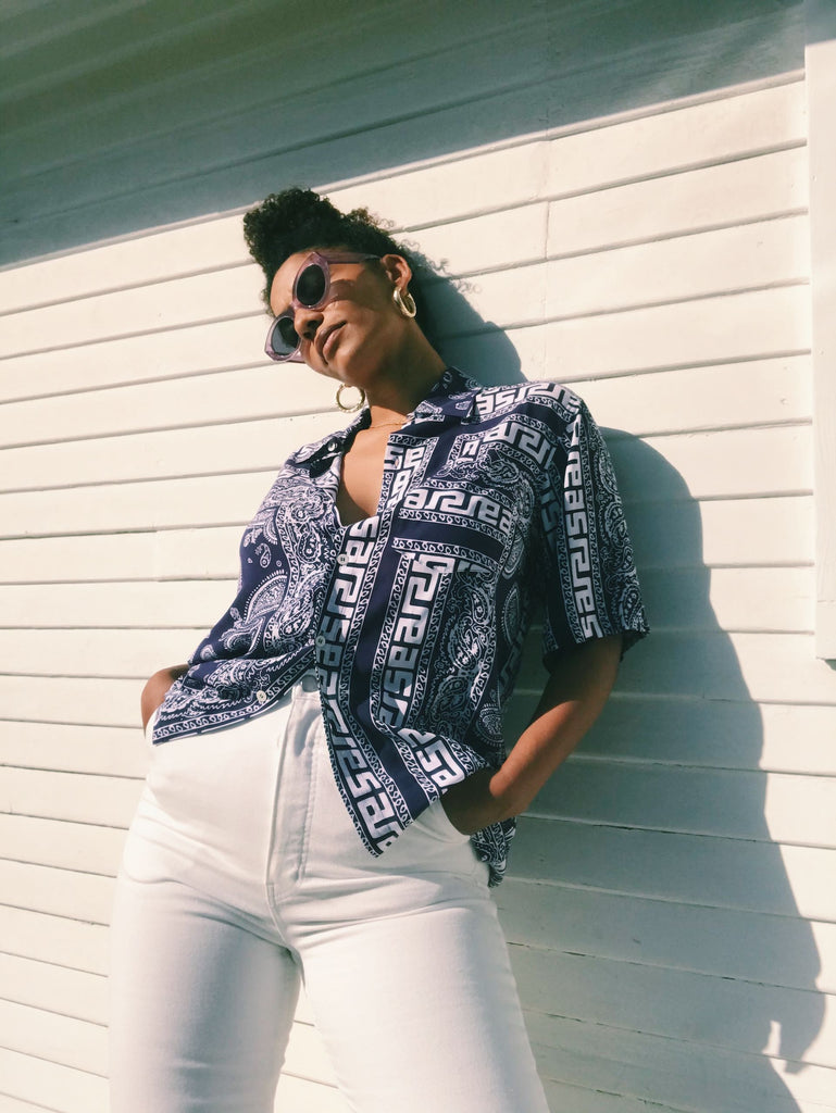 Model stands in front of a paneled white outdoor wall, wearing a printed short sleeve button up and white pants.
