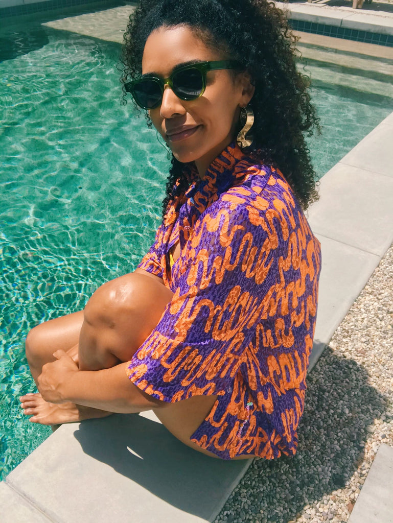 Model sits with her feet in the pool wearing a purple and orange printed and sequined short sleeve button up.