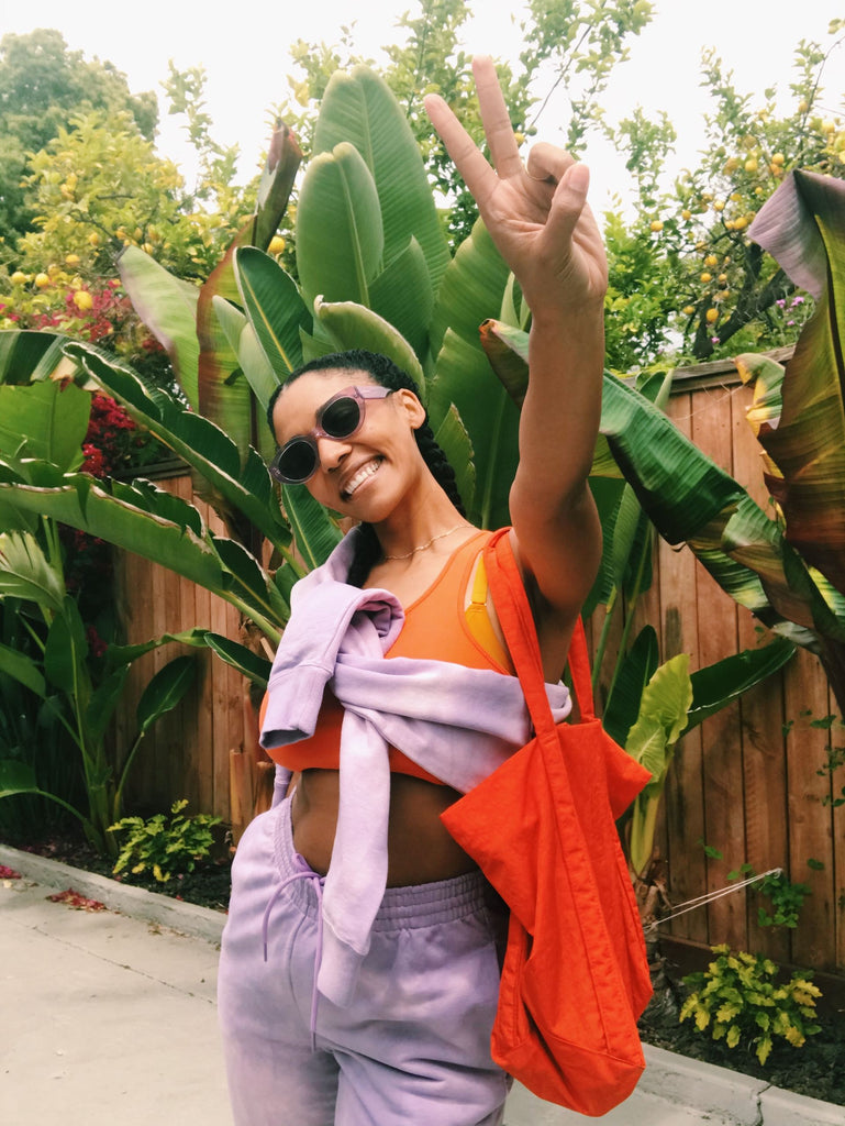 Model stands before green foliage wearing a light purple sweatsuit. The top is tied around her chest over an orange sports bra, and she wears an orange tote bag. She is smiling, wearing sunglasses, and has her left hand holding up a peace sign. 