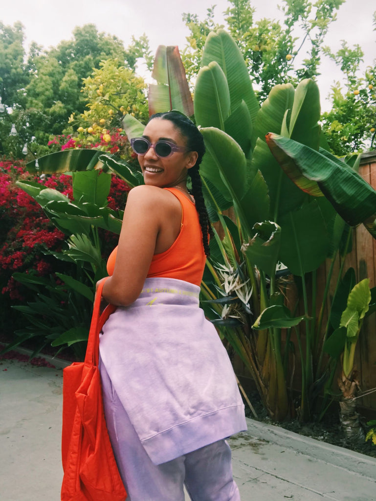 Model stands before green foliage wearing a light purple sweatsuit. The top is tied around her waist over an orange sports bra, and she holds an orange tote bag. She is smiling, wearing sunglasses, and looking over her shoulder.