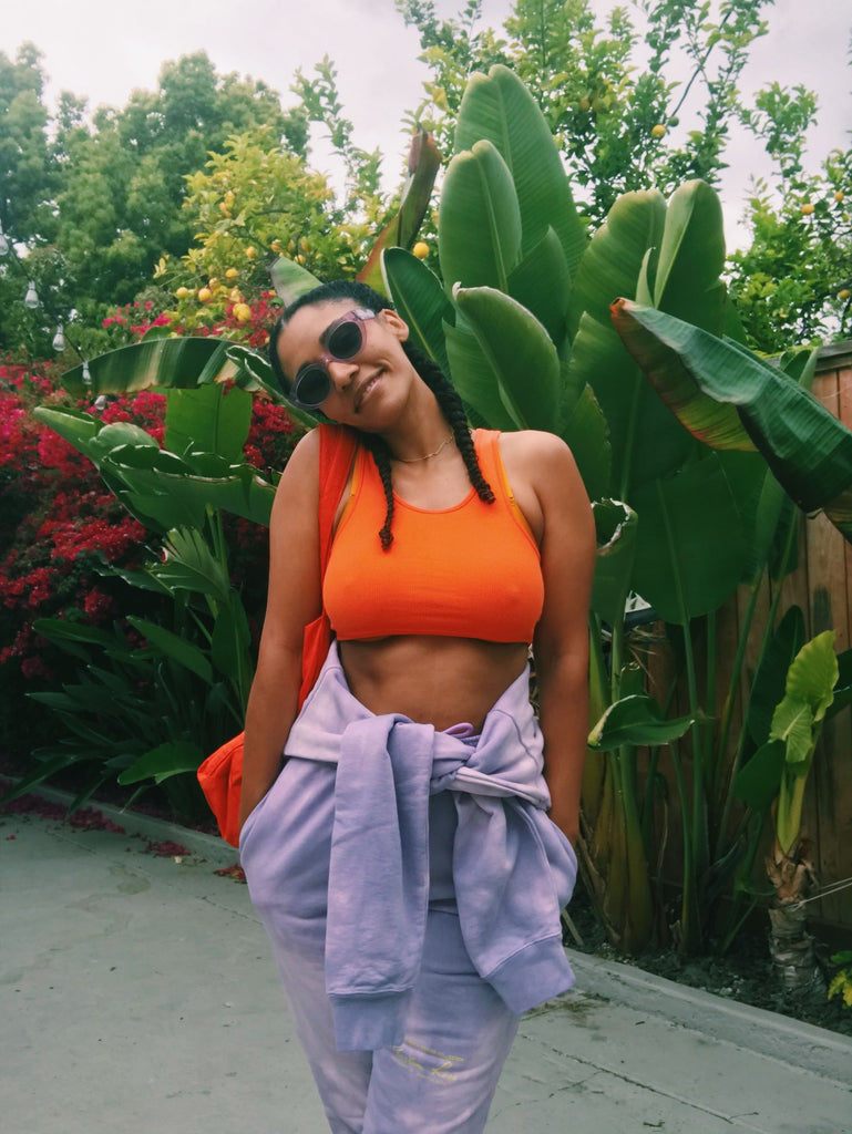Model stands before green foliage wearing a light purple sweatsuit. The top is tied around her waist over an orange sports bra. She is smiling, wearing sunglasses, and has her hands in her pockets.