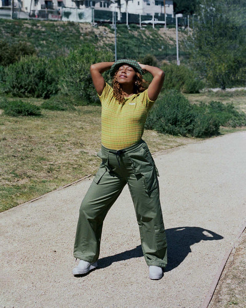 Evelynn standing on a dirt path in a park, holding her hands up behind her head, wearing a yellow check shirt by Stussy and military green pants by Perks and Mini.
