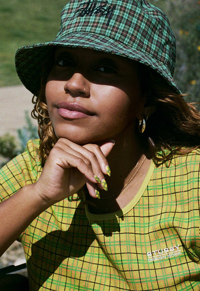A portrait photo of Evelynn, wearing a green plaid hat and yellow checked t-shirt by Stussy, with her chin resting on her hand.