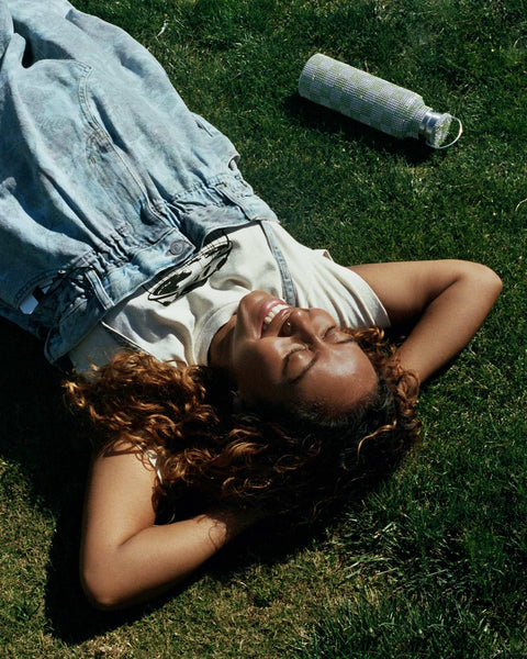 A upside down, angled photo of Evelynn, wearing a neutral tone t-shirt and denim dress by Ganni, laying in the grass on her back, with her arms behind her head.