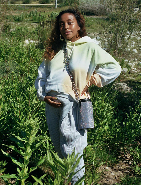 A photo of Evelynn standing amongst grass and greenery, wearing a the-die Collina Strada Sweatshirt, grey Pleats Please pants, and a Kara rhinestone water bottle holder, with her hand under her pregnant belly.