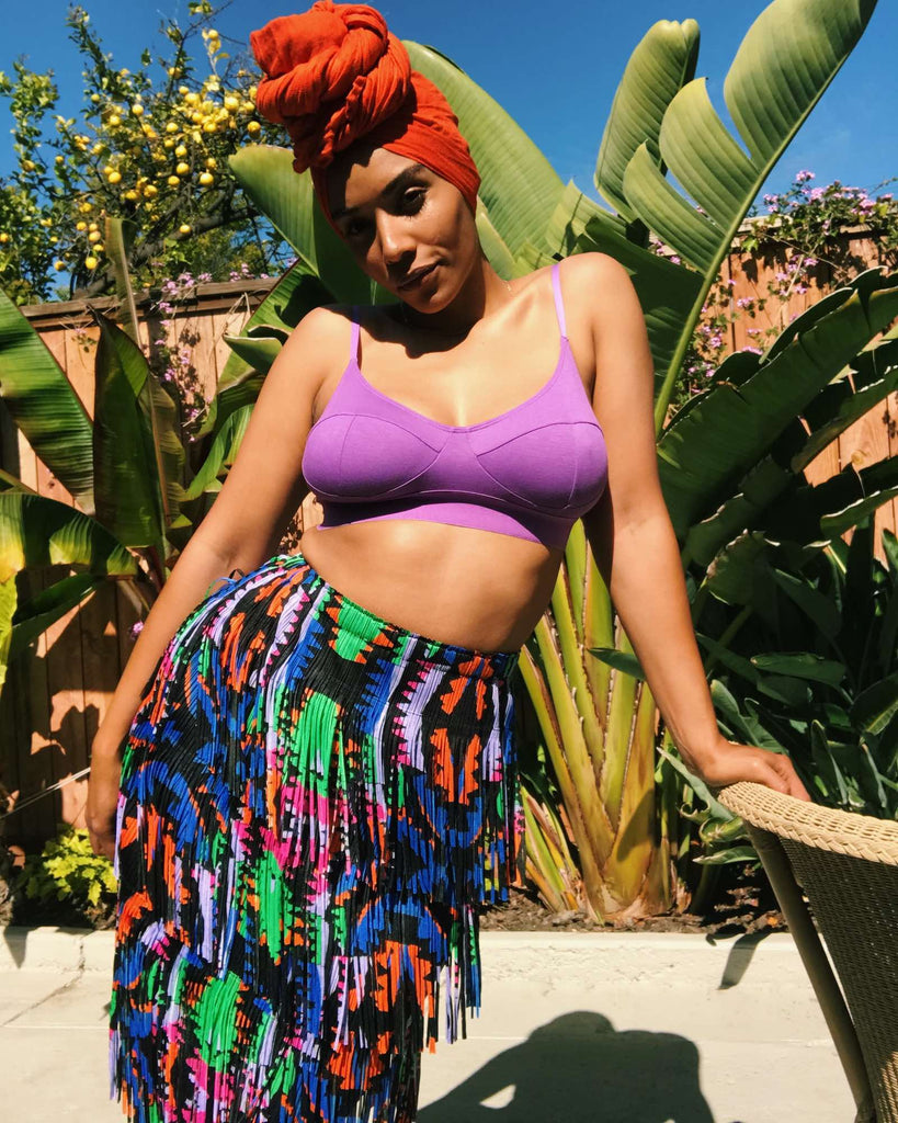 Courtney standing outdoors wearing a purple bra and printed, pleated, fringed skirt in blue, green, orange, and pink.