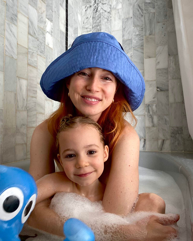 Tiff, wearing a blue hat by Kkco, and Beanie, her daughter, sit in the bath smiling.