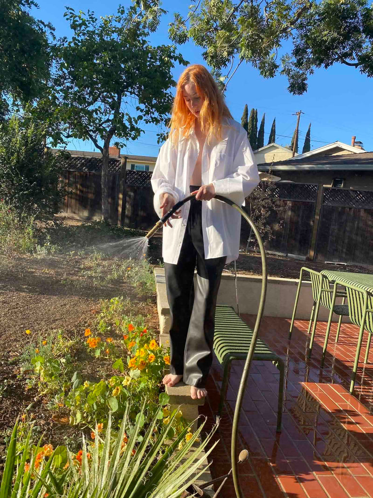Photo of a red-haired woman wearing black leather trousers and a white button up standing and using a hose to water plants.