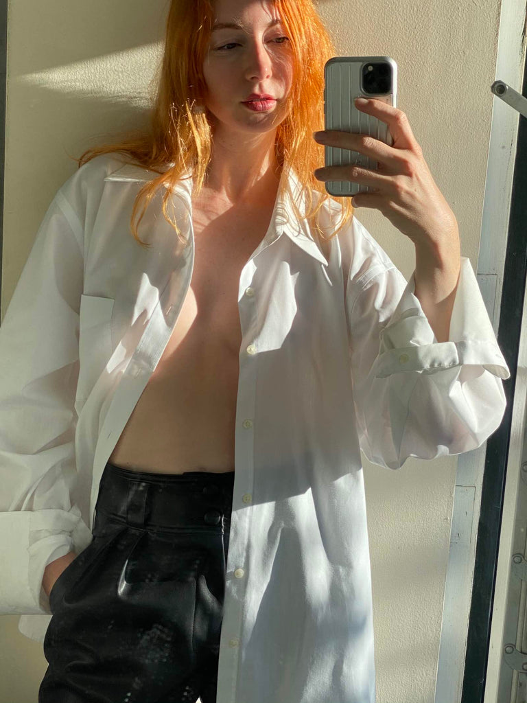 Photo of a red-haired woman taking a mirror selfie wearing leather trousers and a white button down.