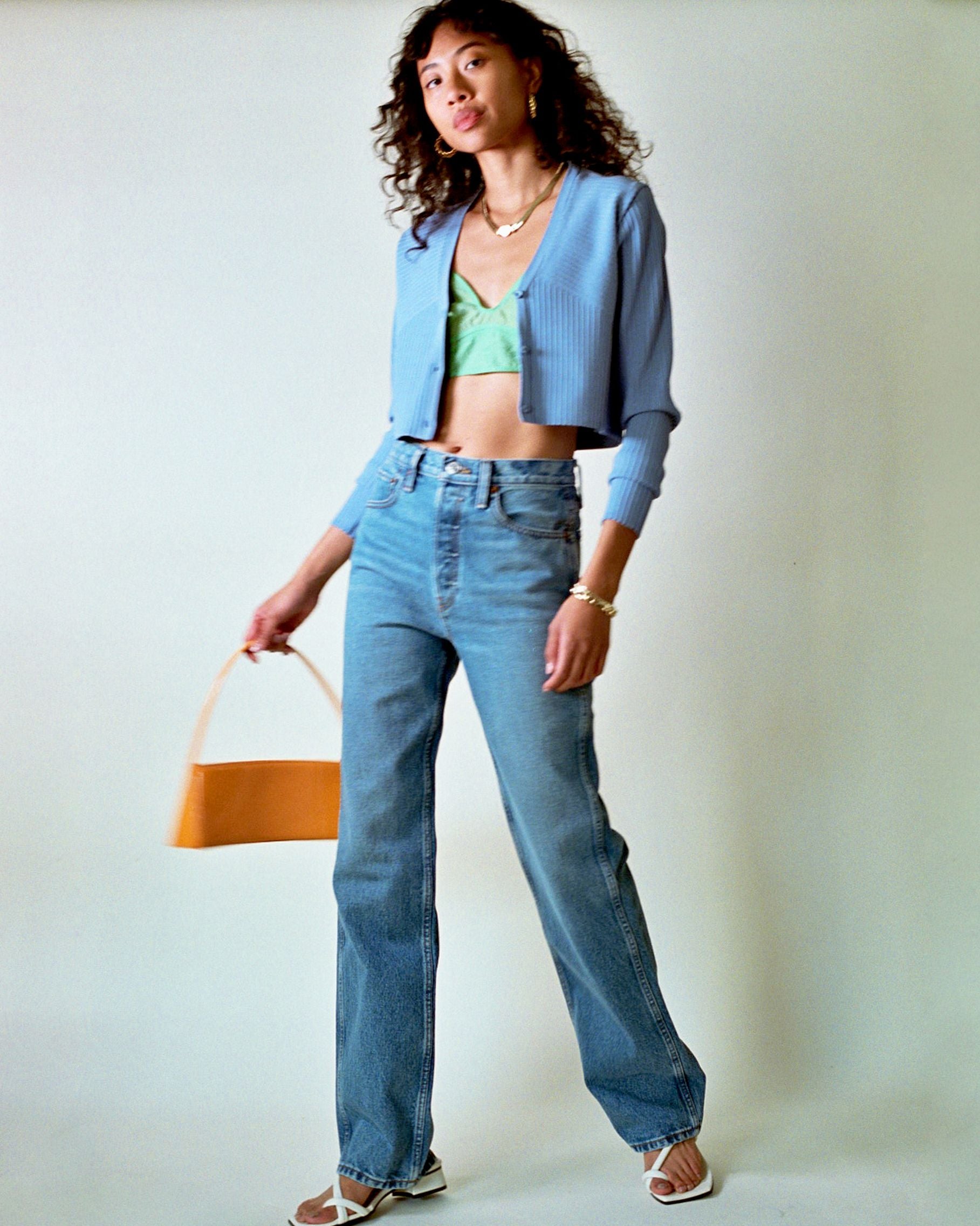 photo of model wearing baggy blue jeans paired with a bright green bra top, light purple knit cardigan and chunky gold jewelry 