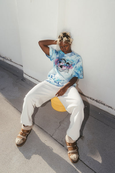 Photo of a model sitting against a wall, wearing a blue tie dye graphic tee from Aries x Pure Beauty x LCD.