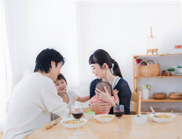 A family sits at a dinner table, mom holds a baby while dad and toddler look on.