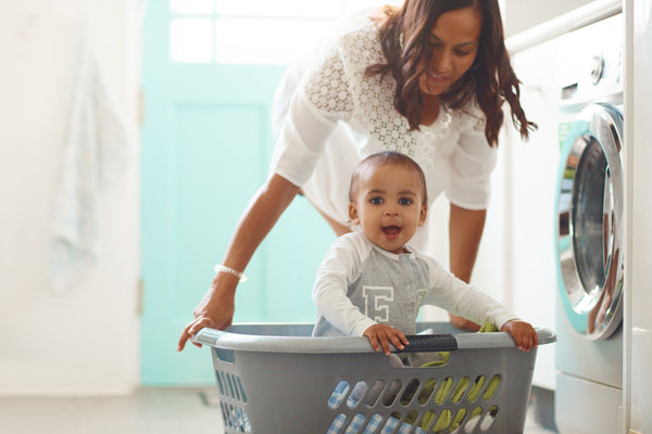 A woman pushes her smiling child in a laundry basket in front of a washing machine.