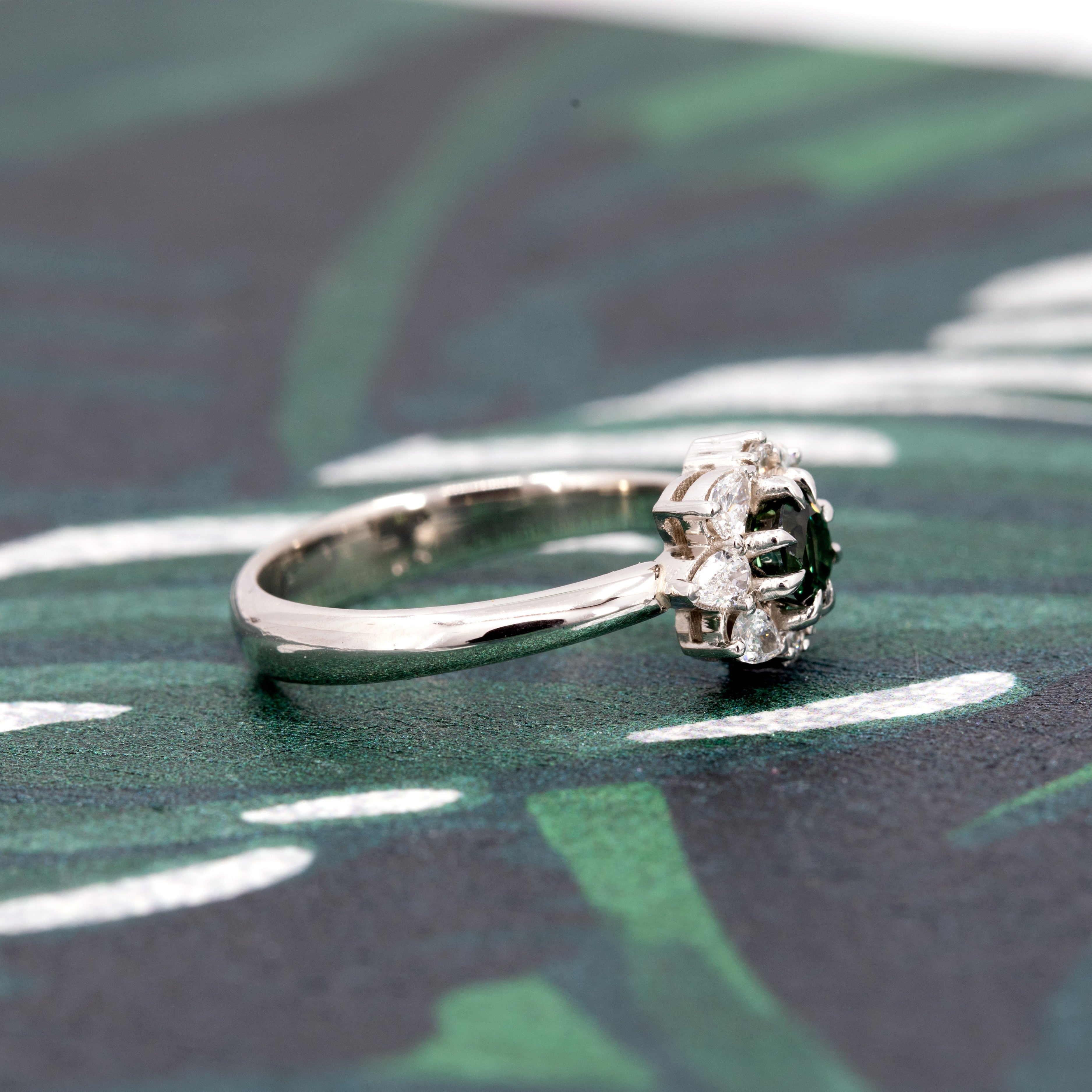 The Deep Forest Glow ring in white gold, displayed on a dark background. The rich green sapphire glows with intensity, framed by a radiant diamond halo for a luminous effect.