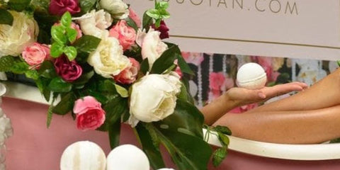 A pink bath surrounded by flowers. A pair of tanned legs are hanging over the side of the bath & a hand is holding a bath bomb. 