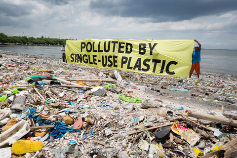 Huge heaps of plastics washed onto the coastline. Two people hold up a large banner that states: polluted by single-use plastic