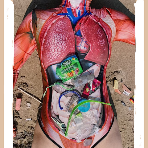 Plastics gathered from a beach walk placed on top of the torso of a person wearing a bodysuit laying on top of the sand in a similar manner to the images of wildlife killed from ingesting plastic. 