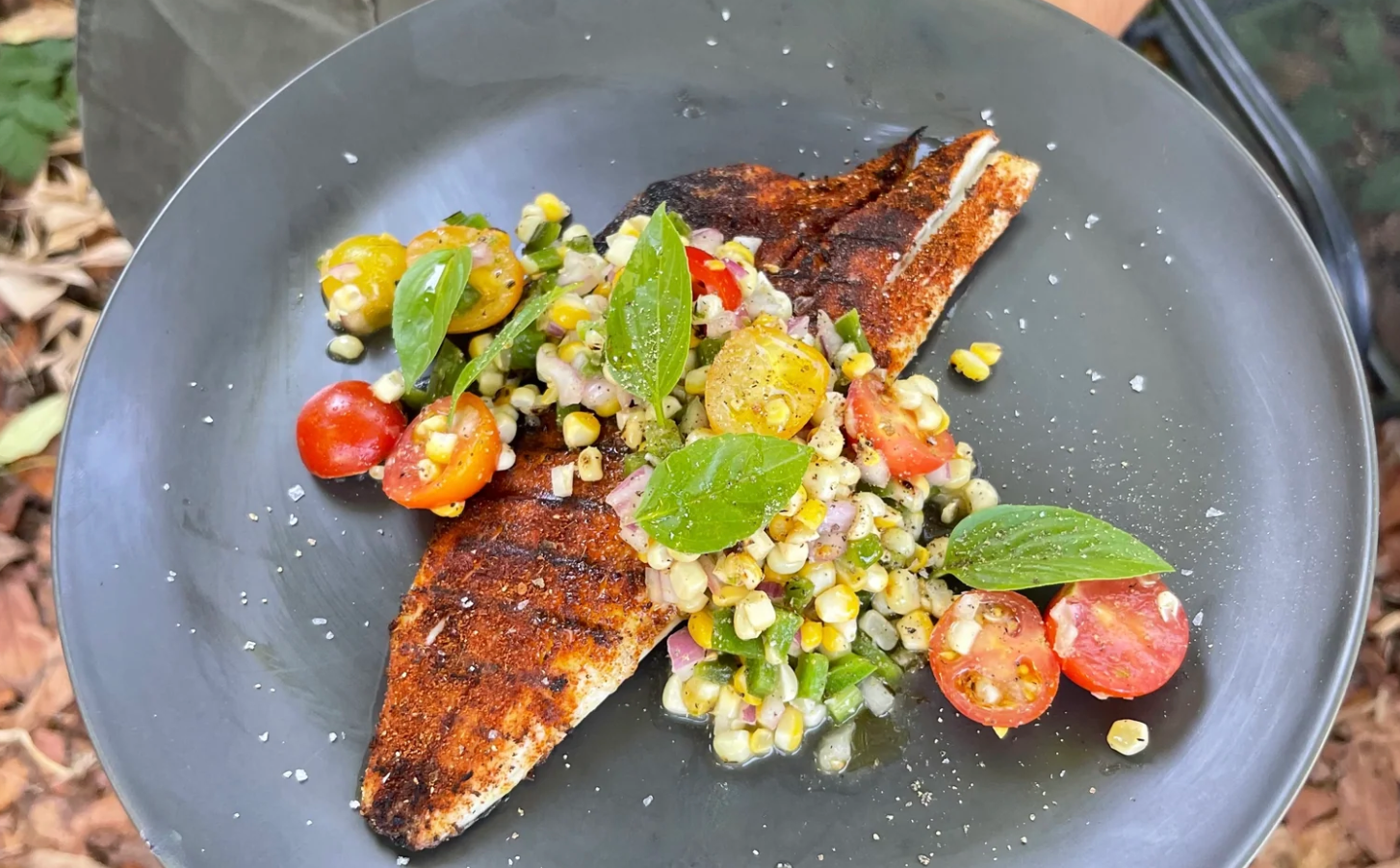 Grilled fish topped with a fresh corn, tomato, and basil salad on a dark gray plate.