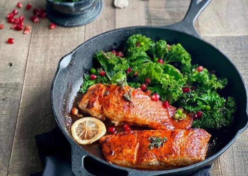 Black pan with cooked salmon and kale on reclaimed wood dining table