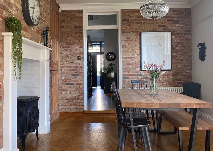 Industrial dining table next to white fireplace and woodburner with exposed brick walls