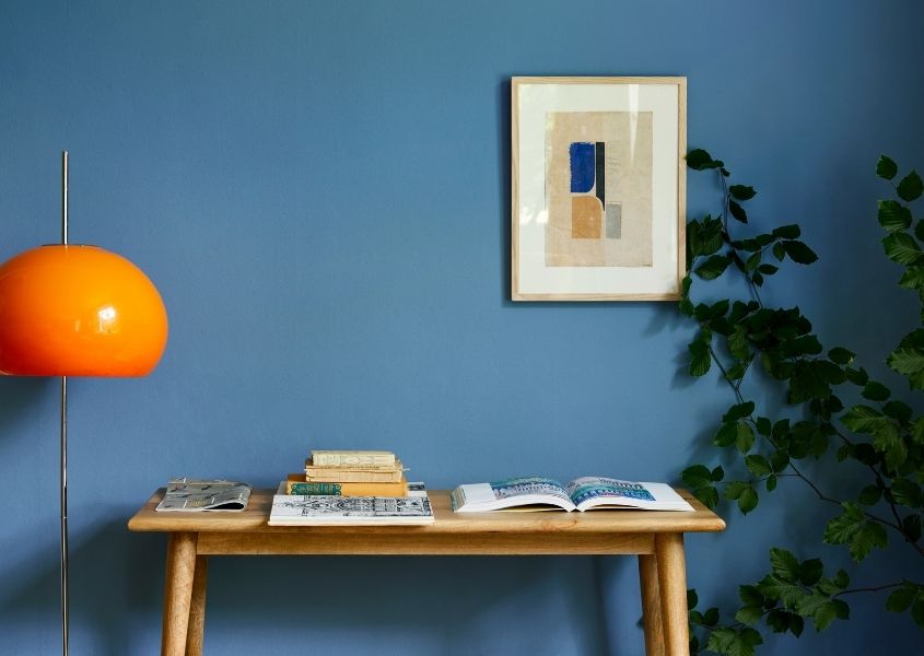 Wooden console table against bright blue painted wall with orange floor lamp and green plant