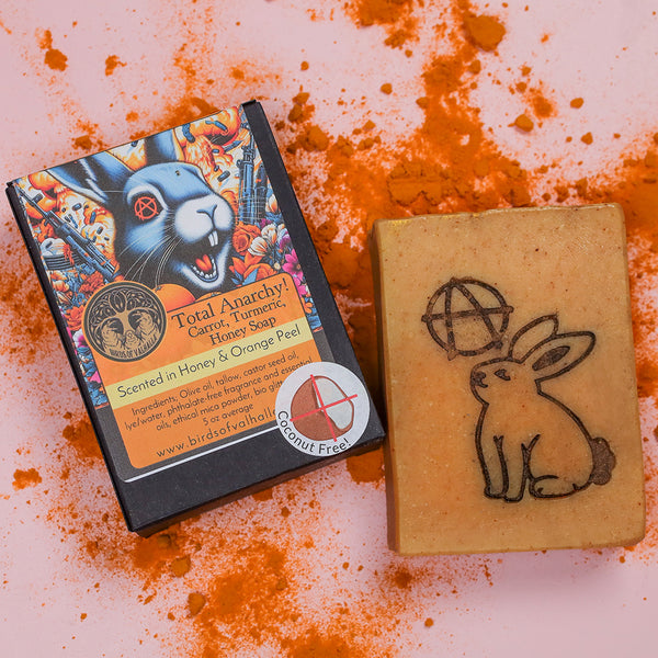 A bar of carrot and turmeric handmade soap, stamped with an anarchy symbol and cute bunny, rests on a chaotic dash of bright orange turmeric.
