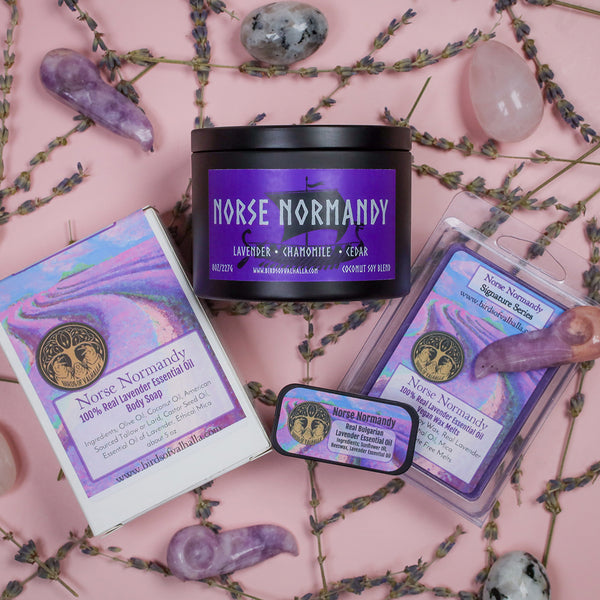 An assortment of items in the Norse Normandy fragrance- a candle, wax melts, solid cologne, and handmade soap, as well as a variety of crystals- on a pink background surrounded by real lavender.