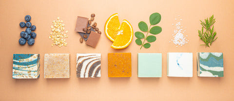 7 soaps in a row, above each one displays an ingredient that is implied to be a food ingredient in the soap. From left to right the ingredients are: blueberries, oats, chocolate and cacao, orange slices, mint, salt, and rosemary.