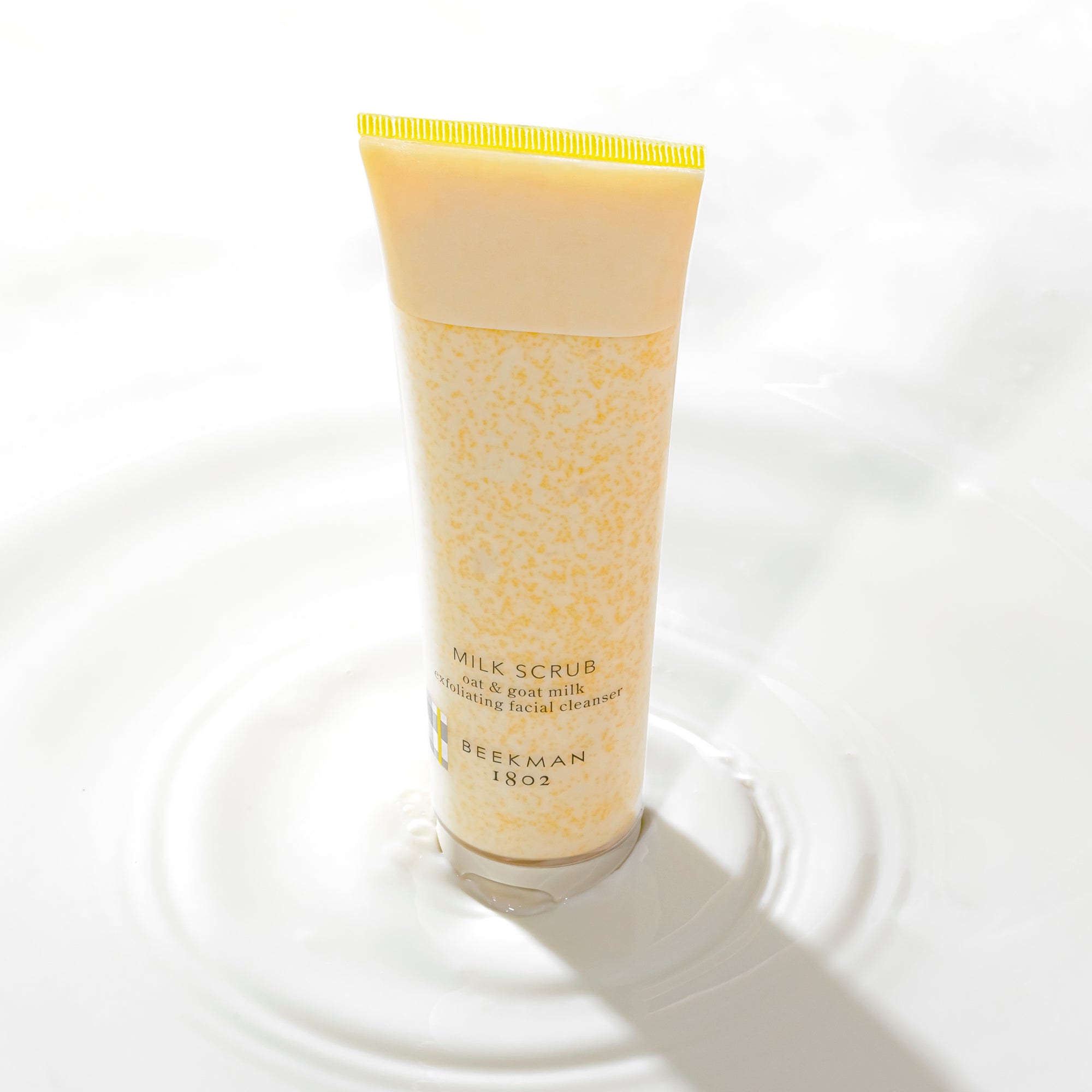 Bottle of Beekman 1802's Milk Scrub Facial Cleanser surrounded by a pool of milk and causing a ripple in the milk.