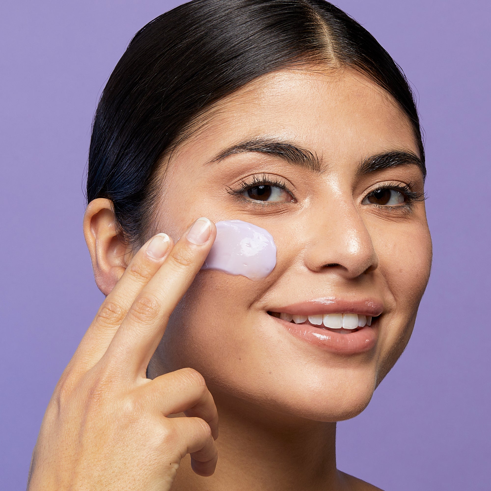 Head shot of model applying beekman 1802's purple midnight milk mask onto her face using her fingers, on a purple background.