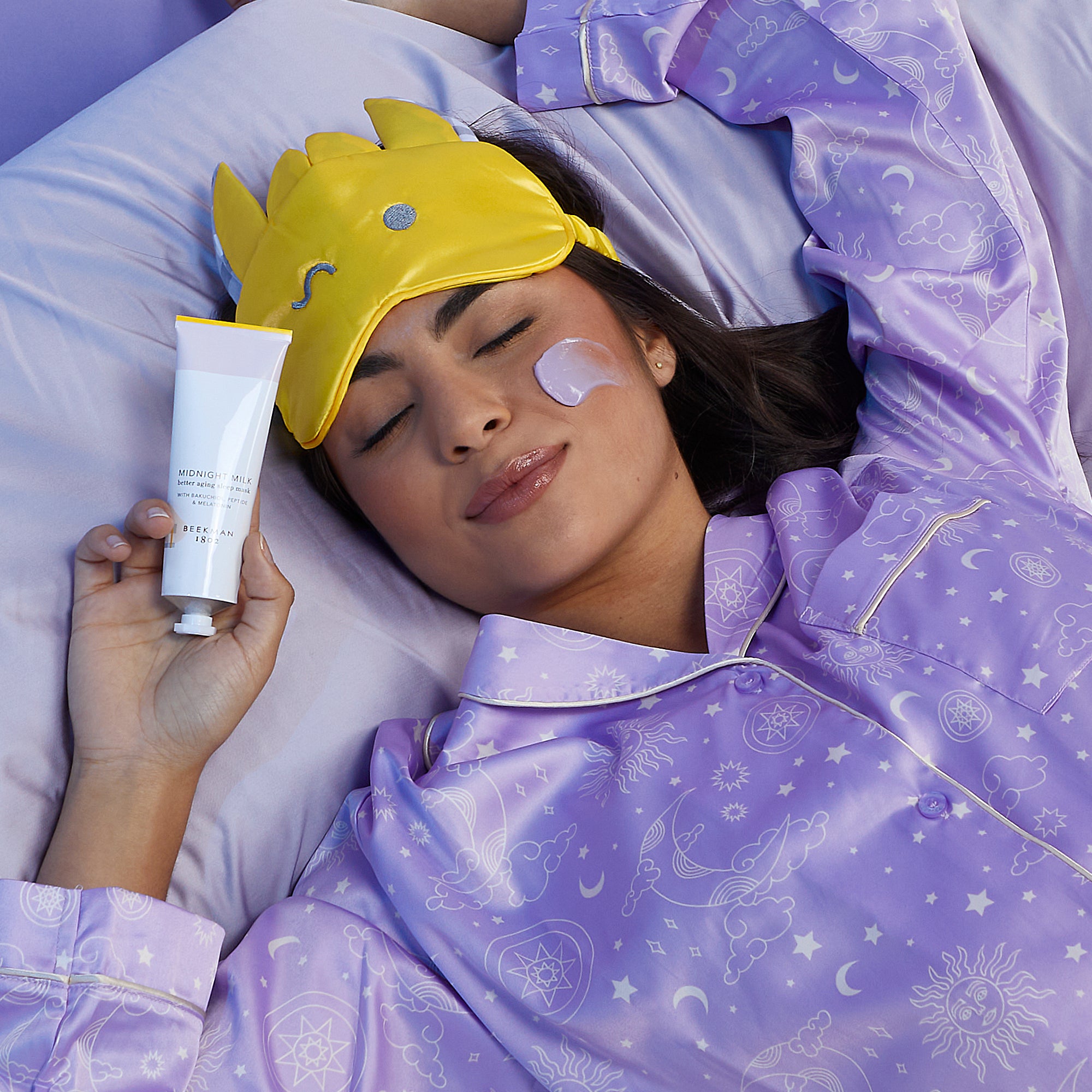 Model wearing purple pajamas and a yellow eye mask and laying on her bed while holding a tube of Beekman 1802's Midnight Milk Sleep Mask up to her face with her eyes closed, and the purple mask product swatch on the side of her cheek.