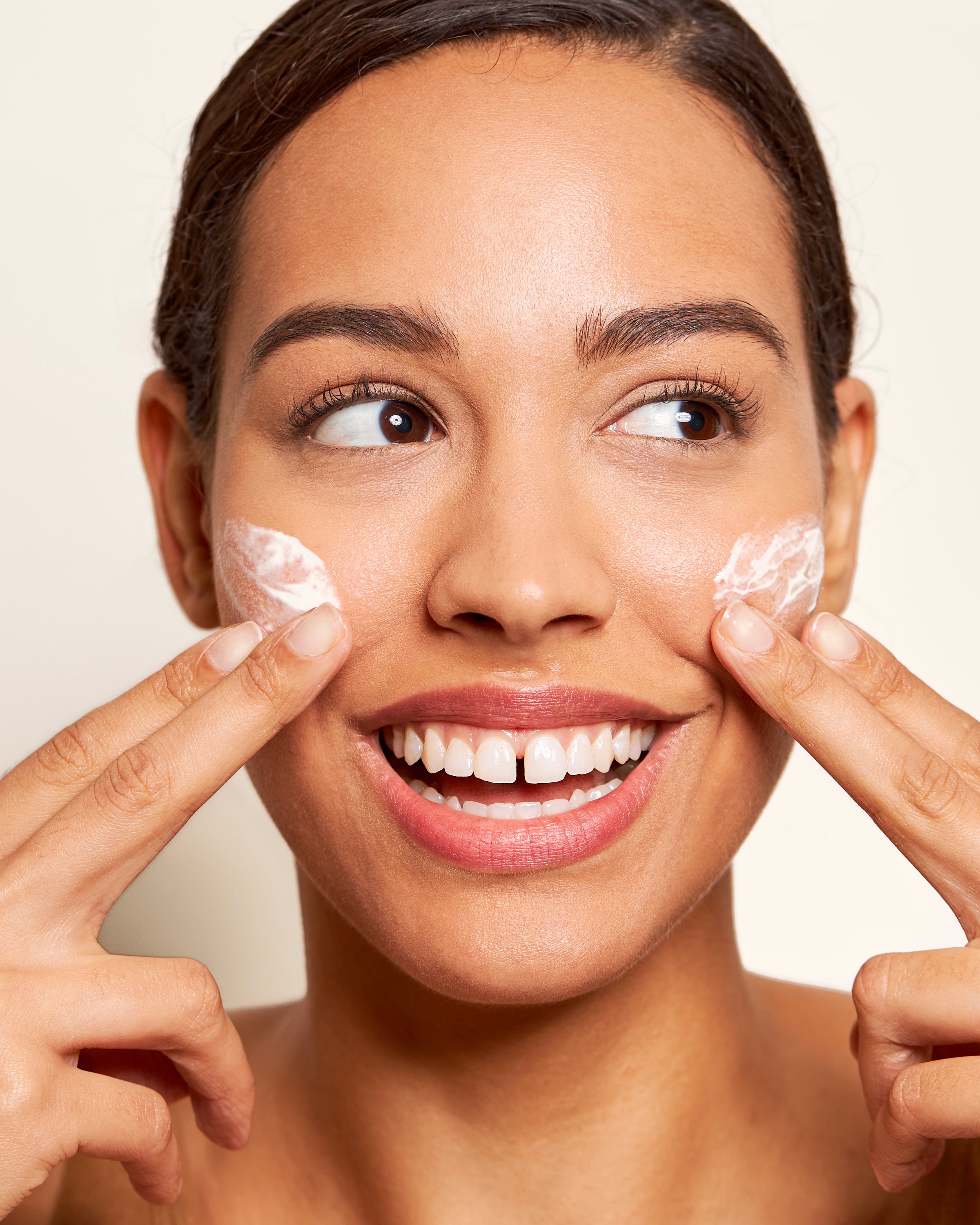 Headshot of model applying Beekman 1802's Bloom Cream Daily Moisturizer to both sides of her cheeks with both of her hands, while looking off to the side and smiling.