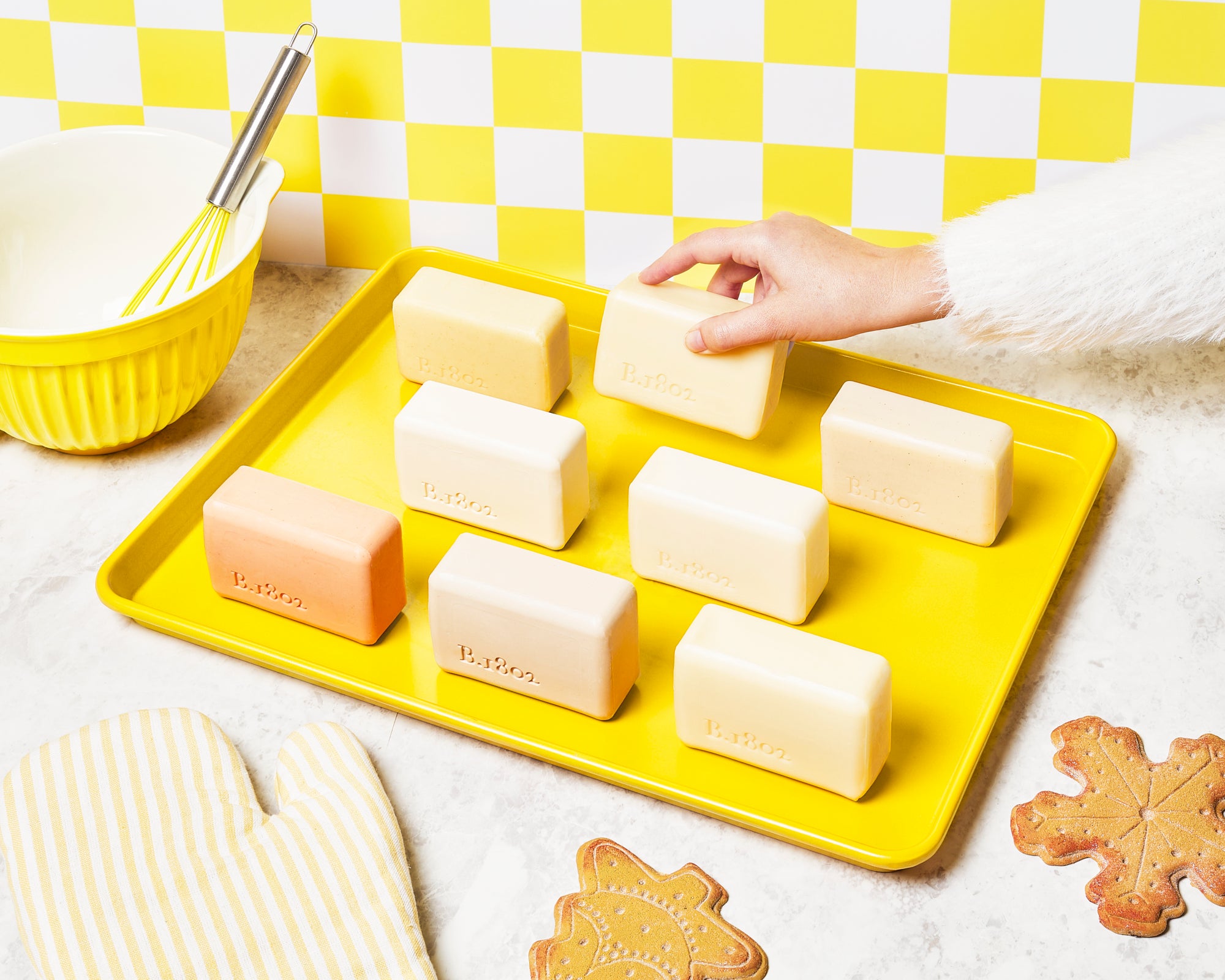 8 unwrapped Beekman 1802 goat milk bar soaps all lined up on a yellow baking tray, with a hand coming from the right side of the image picking up a bar soap, with the tray surrounded by cookies, oven mitts, a mixing bowl and whisk, on a yellow and white checkered background.