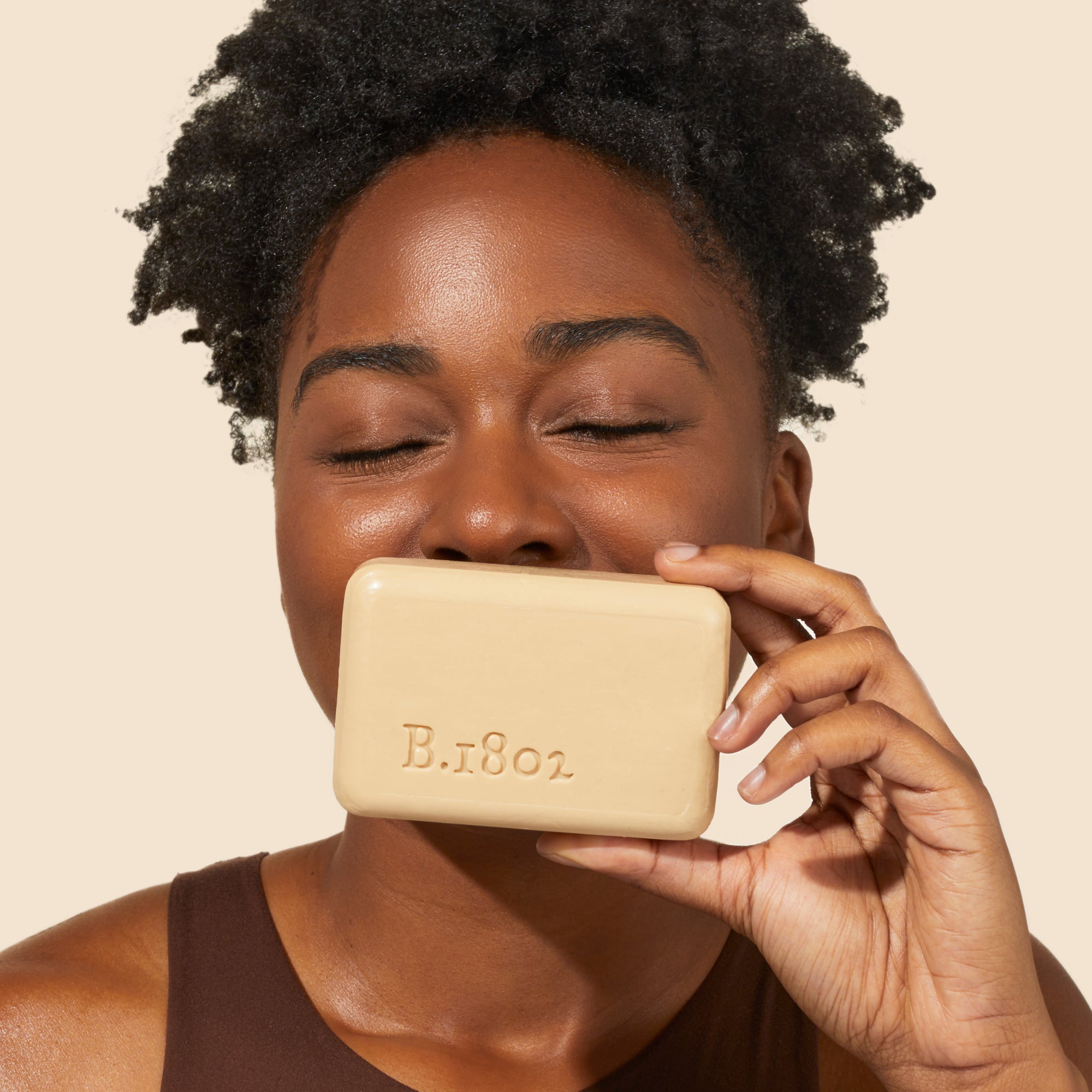 Up close shot of model holding up an unwrapped Beekman 1802 goat milk bar soap to her nose and smelling it with her eyes closed, on a cream colored background.