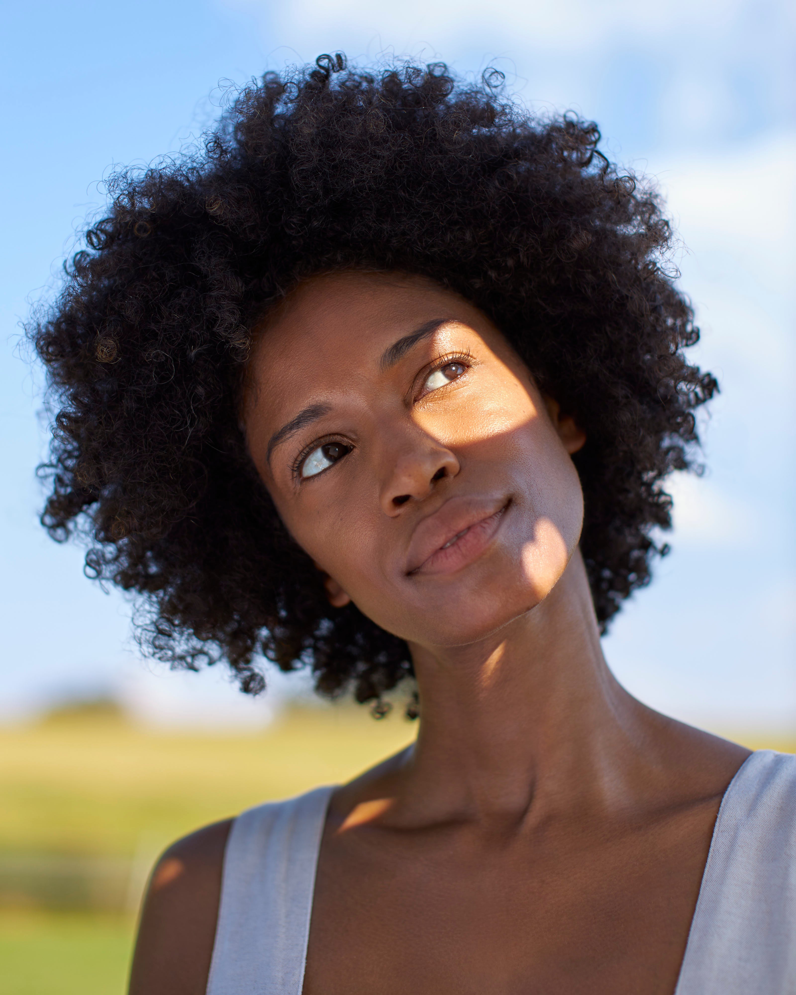 Head shot of model looking off to the side with the sun shining on her face, and blue sky and outside in the background.